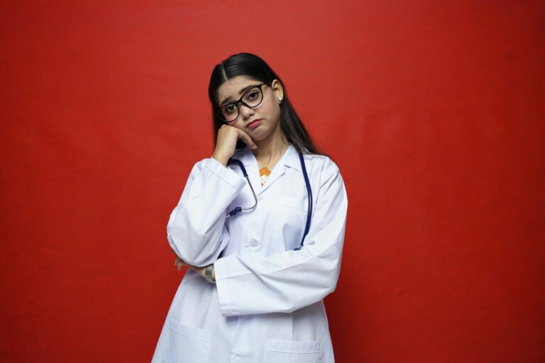 A female hispanic doctor in a white lab coat, with a stethoscope and dark rimmed glasses. Her head is tiled to the right, resting in her right hand. She has a sad expression.
