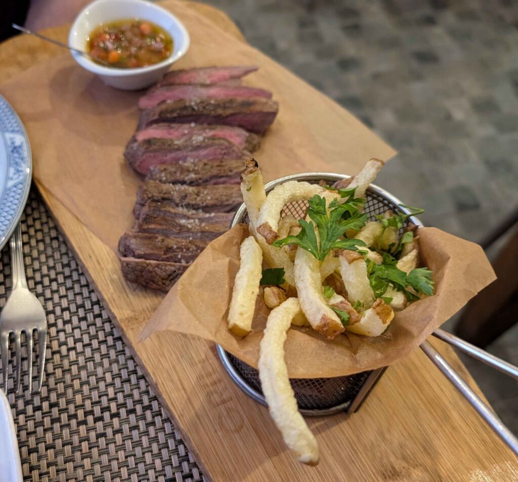 A serving board with grilled steak and a small basket of french fries.