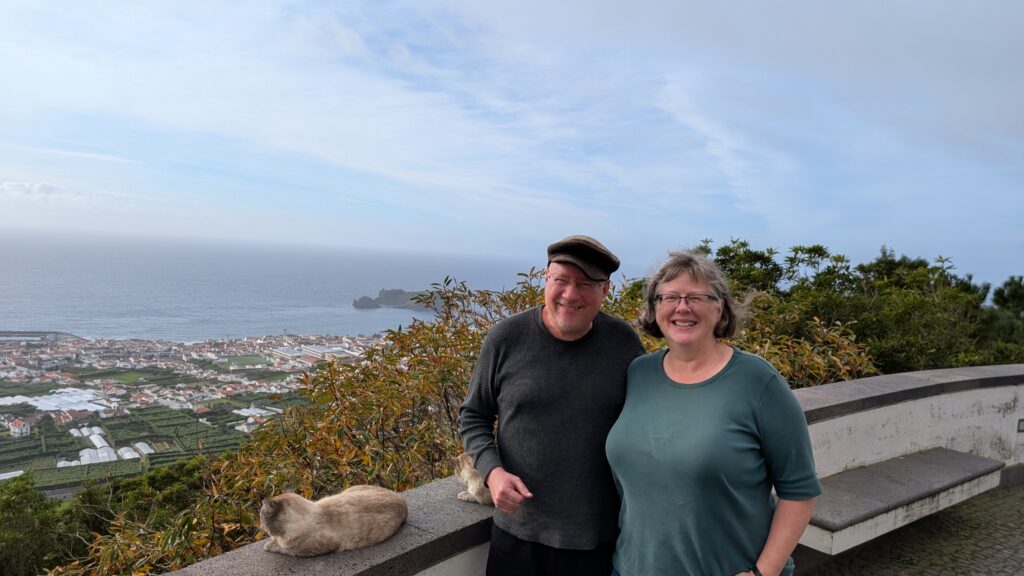 The author and their spouse on a porch overlooking the town of Furnas.  There is a cat on the ledge of the porch.